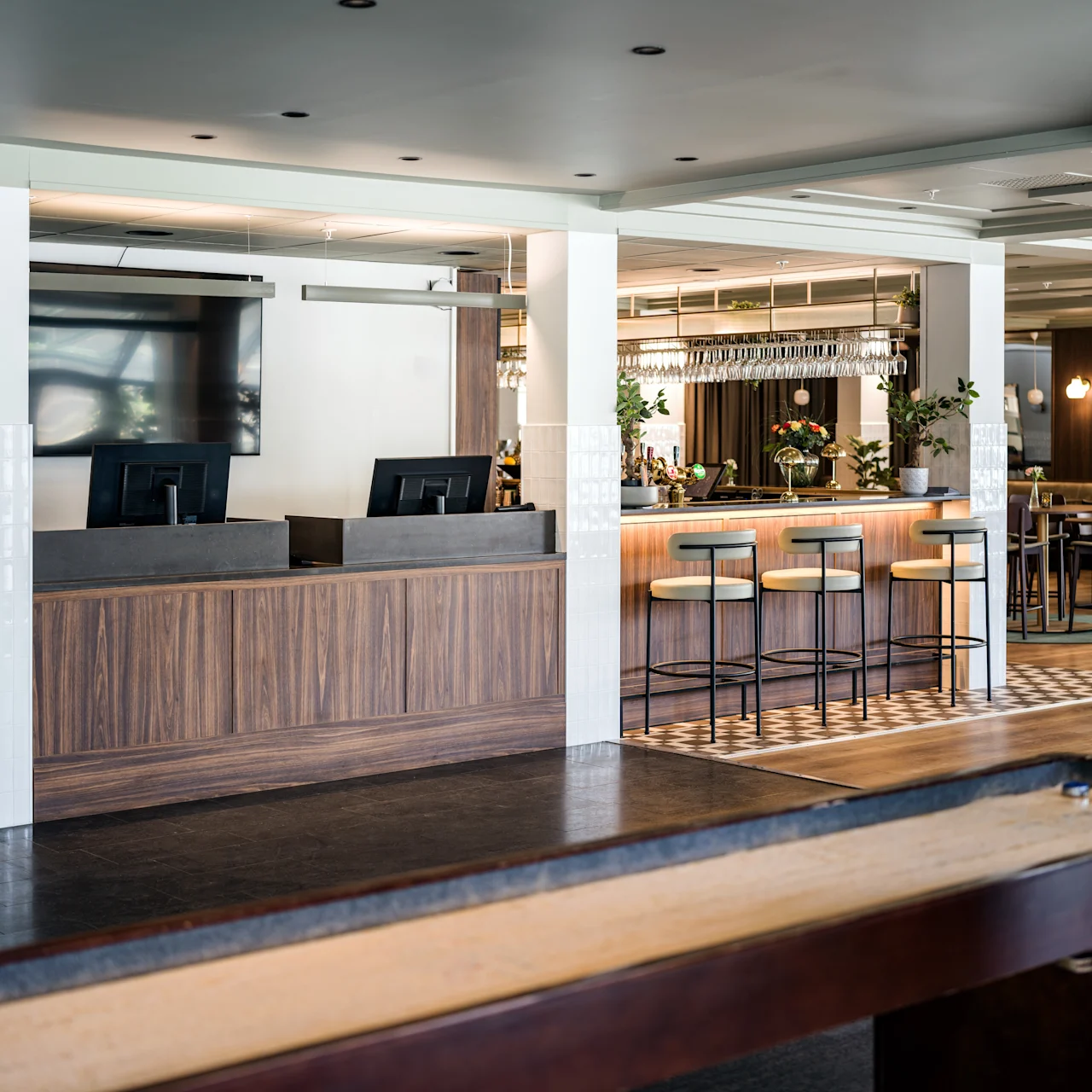Reception desk stands empty beside a modern bar with high stools, surrounded by indoor plants and ambient lighting. A shuffleboard table occupies the foreground.