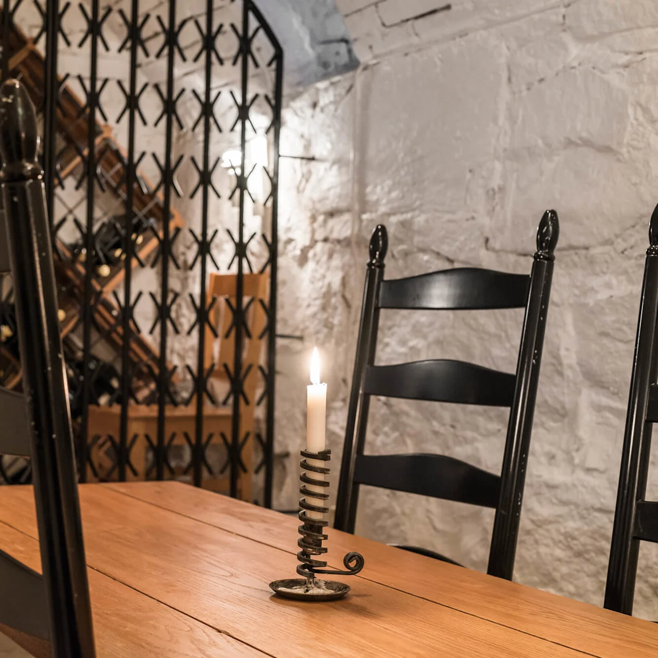 A wooden dining table with black chairs and a lit candle, set against a wine rack and white brick wall.