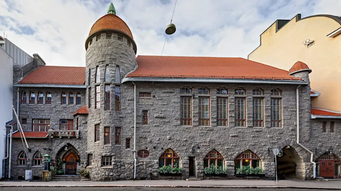 En historisk steinbygning har et sylindrisk tårn med rødt tegltak, plassert i en urban gatekontekst under en skyet himmel.