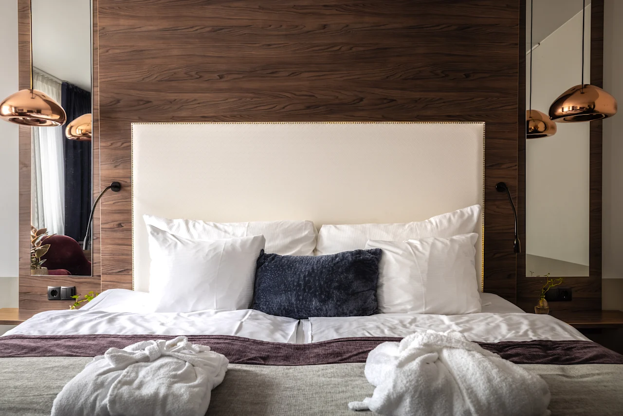 Mukava hotellihuone, jossa pehmeä sänky on täynnä tyynyjä ja viitat on nostettu esille. Tyylikkäät lamput valaisevat tilaa, ja puuelementit lisäävät lämpöä ja eleganssia.