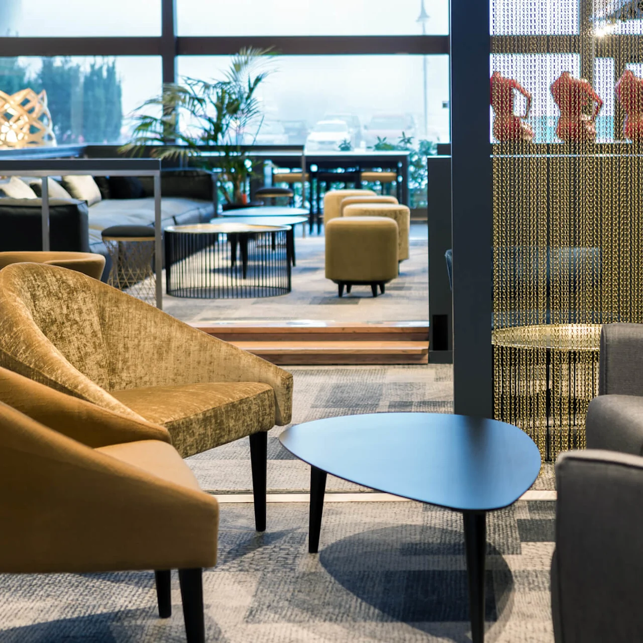 Cozy seating area with various chairs and a triangular table; gold-fringed curtains partially divide the space. Background features sofas, plants, and a large window with foggy exterior view.