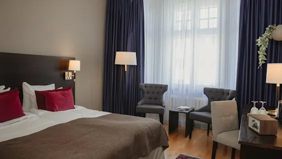 Dobbeltseng med brun dyne og røde puder i hotelværelse. To grå stole vender mod et lille bord ved et vindue med blå gardiner. Et skrivebord har en lampe og dekoration.