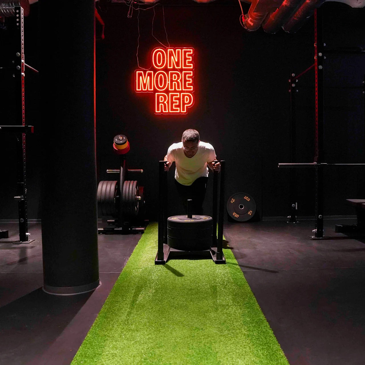 Mann skyver vektet slede på grønt underlag i treningsstudio. "ONE MORE REP" i neonlys på svart vegg. Omgivende utstyr inkluderer benker og vekter.