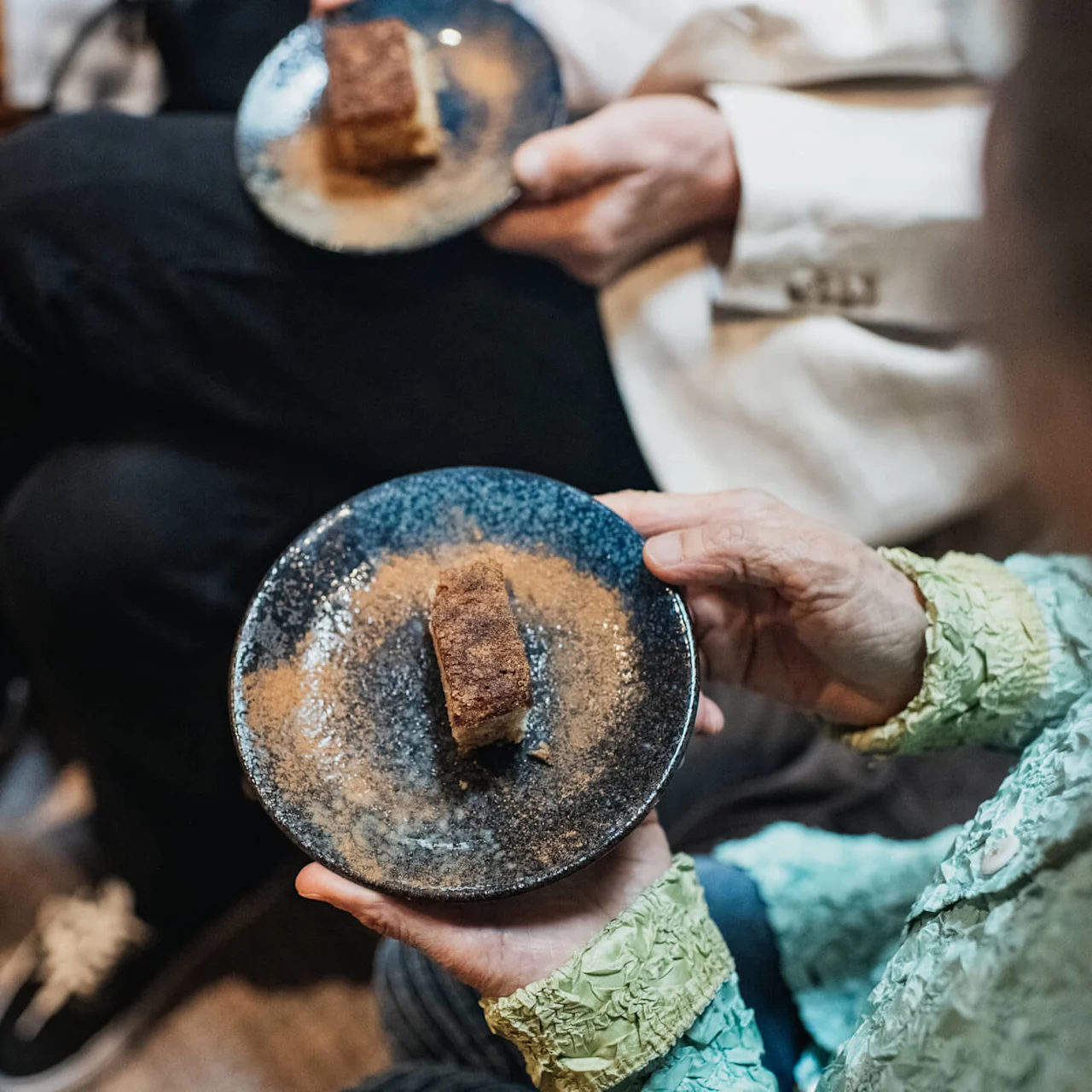 Two people holding plates with a piece of cake, partially sprinkled with sugar, in a cozy indoor setting.