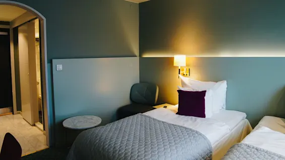 Ett hotellrum med två prydligt bäddade sängar, lila kuddar och takbelysning. Rummet har gröna väggar, ett litet runt bord och en öppen badrumsdörr.