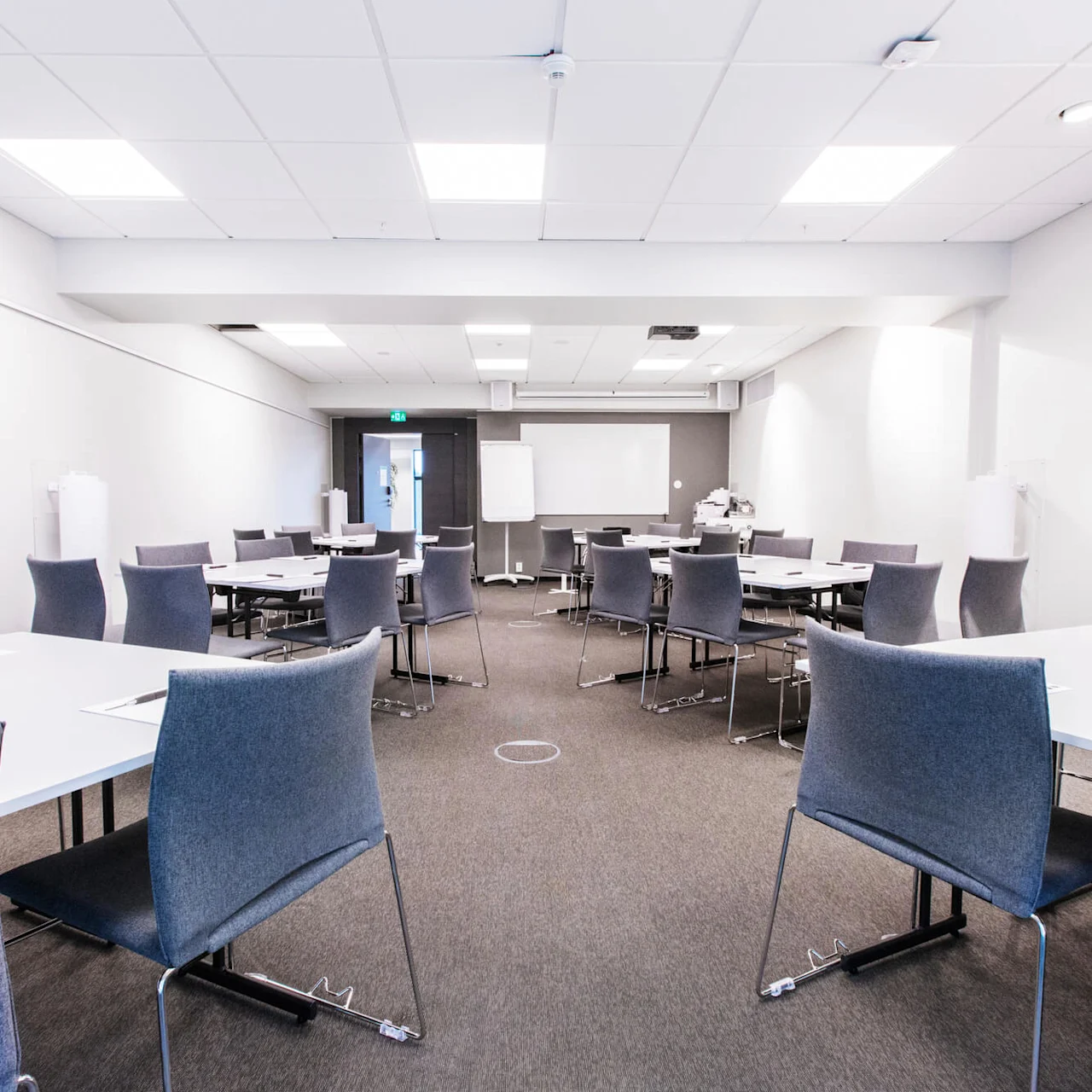 Bord med stolar arrangerade i ett ljust konferensrum; pennor och block på borden i en heltäckningsmattsmiljö med whiteboard och blädderblockställ framme.