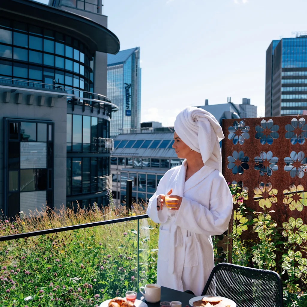 En person i badekåpe nyter frokost på en grønn balkong med utsikt over en bysilhuett en solrik dag.