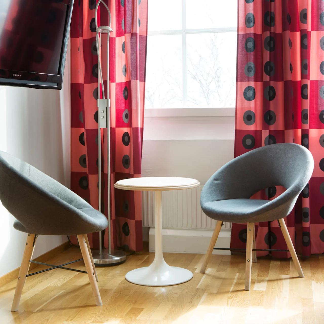 Two gray chairs surround a small white table in front of a window with red curtains featuring black circular patterns. A floor lamp stands nearby, and a TV is mounted on the wall.