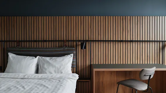 Bed with white linens beside a wooden desk, situated against a wooden slat wall, and accompanied by a modern gray chair in a minimalist, contemporary room.