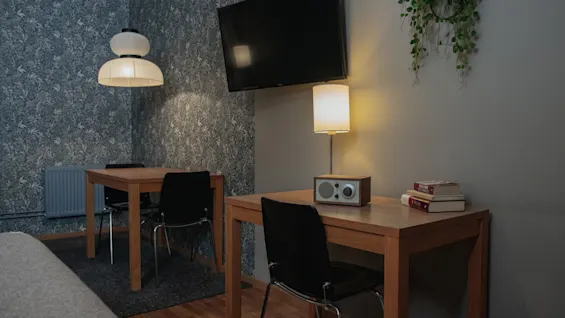 A wooden desk with a table lamp and radio is in a room with a wall-mounted TV. Another desk and a hanging light are in the background with patterned wallpaper.