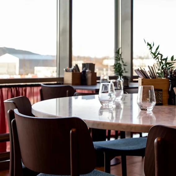 Rundt spisebord omgitt av trestoler, satt med glass. Ligger i en lys restaurant med store vinduer med utsikt over fjellene og røde gardiner under vinduene.