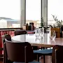 Rundt spisebord omgitt av trestoler, satt med glass. Ligger i en lys restaurant med store vinduer med utsikt over fjellene og røde gardiner under vinduene.