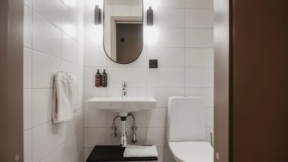 Ett rent, modernt badrum med vita kaklade väggar, ett handfat med toalettartiklar, en spegel och en toalett.