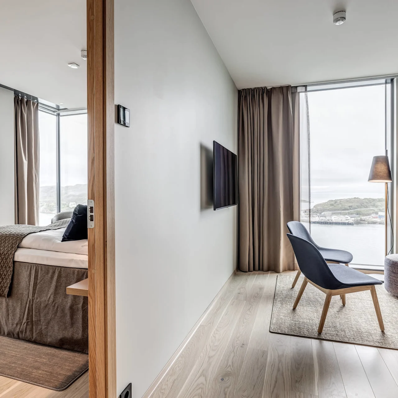 A cozy hotel room with a bed and TV on one side, two chairs and a lamp near large windows overlooking a waterfront on the other side.