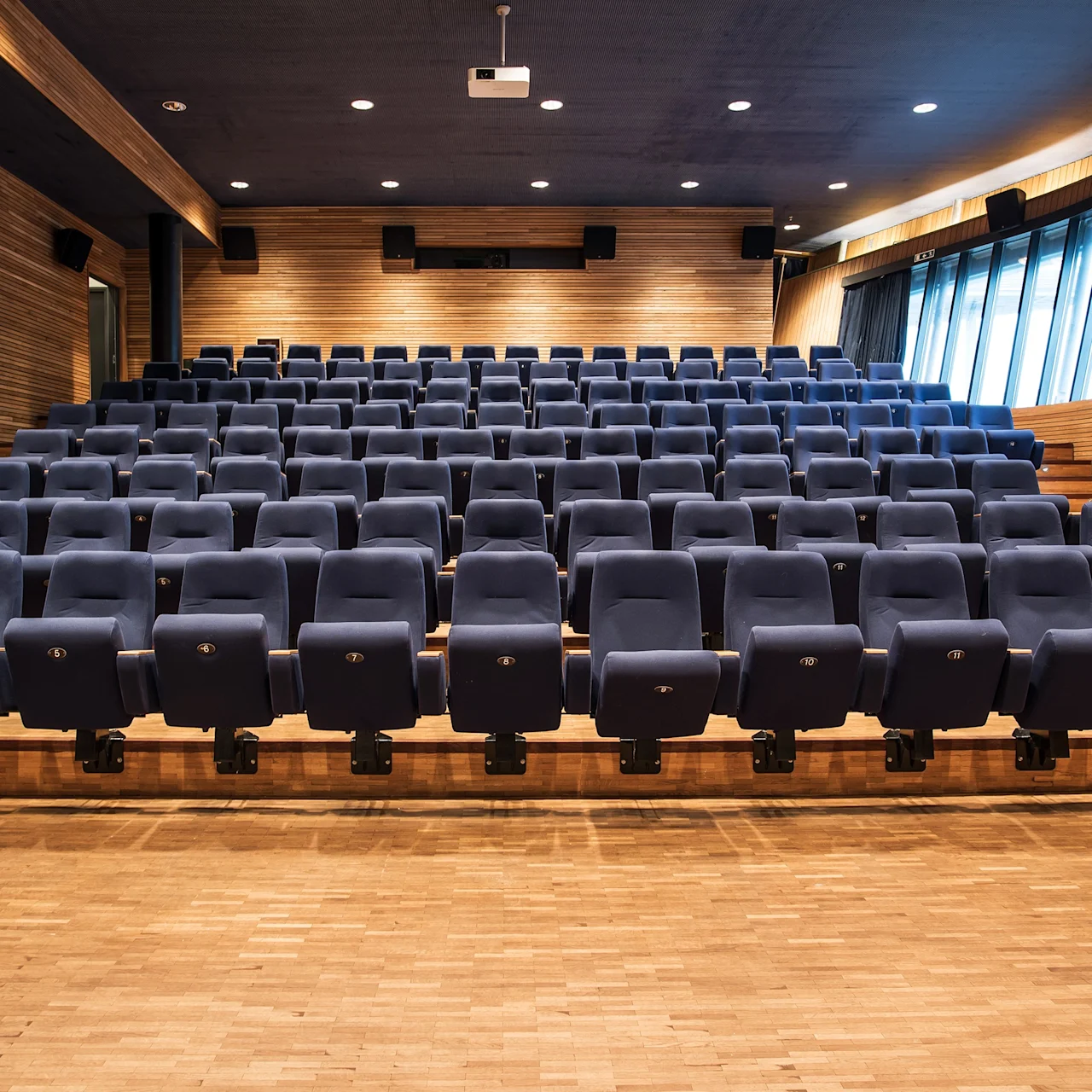Rader av tomme blå teaterseter vender mot en trescene i et auditorium, med trepanelvegger og en projektor montert i taket.
