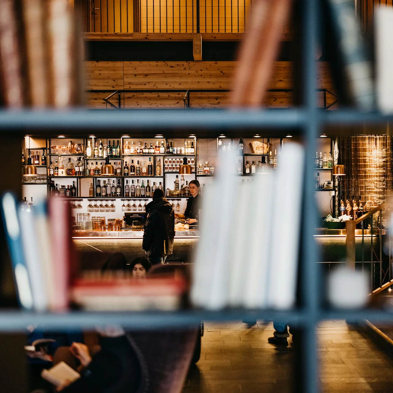 A cozy bar scene viewed through shelves with blurry foreground, patrons interacting, warm lighting, and a wooden interior. Copperhill Mountain Lodge hotel in Are. 