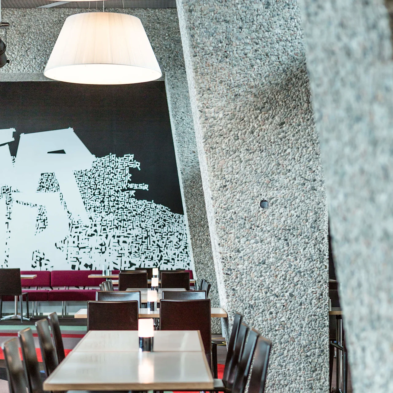 Tables are lined with chairs and lamps, set neatly in a modern dining area surrounded by textured concrete walls and artistic wall decoration with scattered typography.