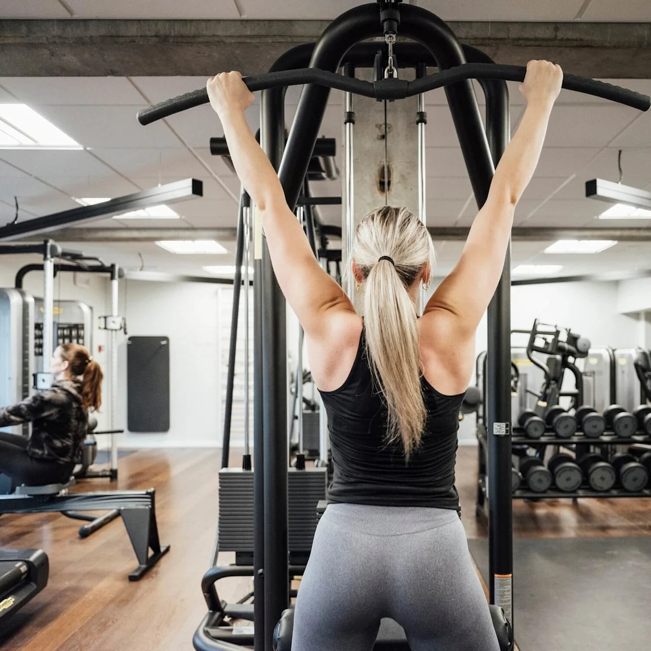 En kvinde laver en lat pulldown-øvelse i et motionscenter, omgivet af forskelligt udstyr og andre mennesker, der træner.
