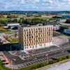 Ett högt, rektangulärt hotell med namnet "The Urban House" står på en stor parkeringsplats, omgiven av ett industriellt landskap med spridda lager och gröna fält under en delvis molnig himmel.