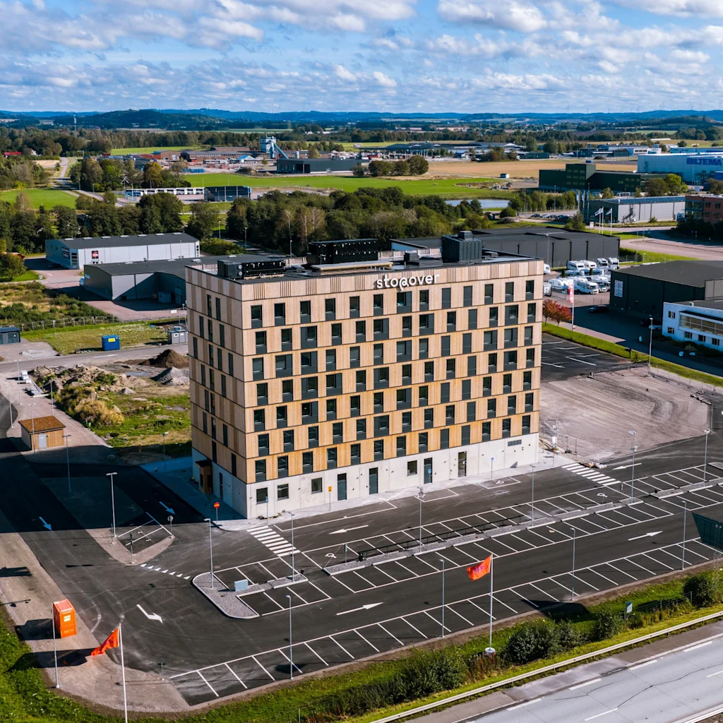 Ett högt, rektangulärt hotell med namnet "The Urban House" står på en stor parkeringsplats, omgiven av ett industriellt landskap med spridda lager och gröna fält under en delvis molnig himmel.