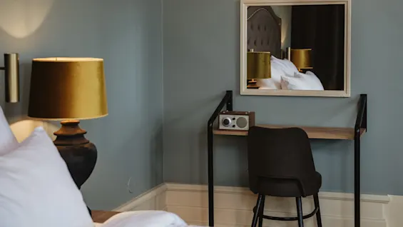 A table with a radio sits beneath a mirror reflecting a bed and a lamp, in a neatly arranged bedroom with gray walls.