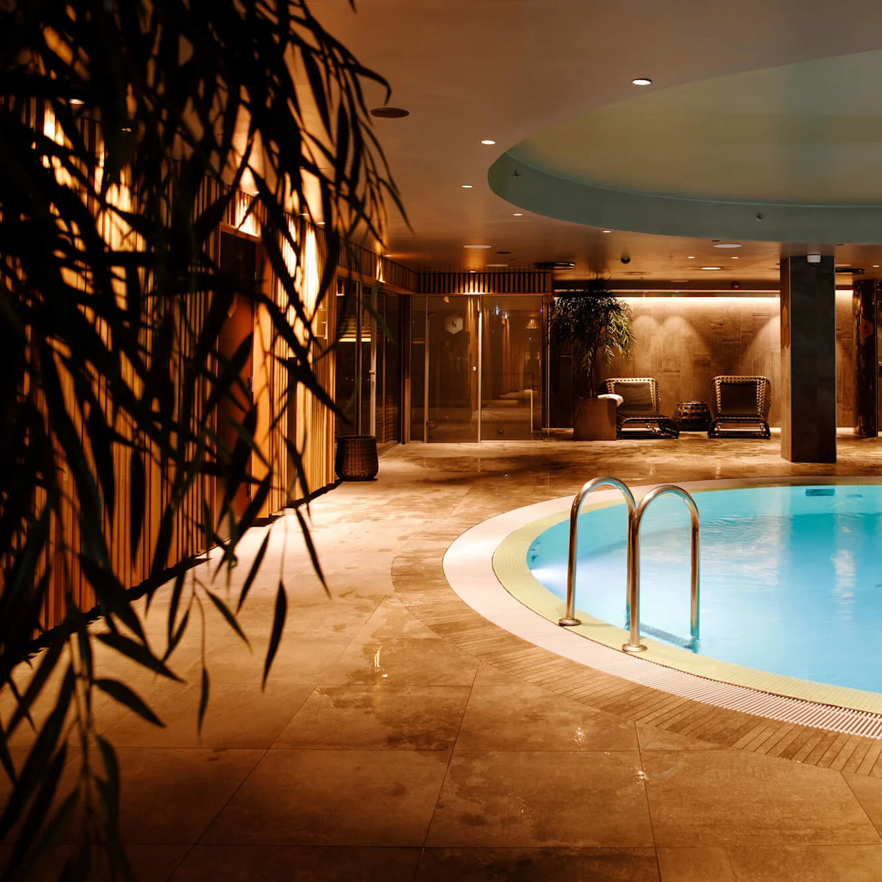 A round indoor pool with a central pillar, surrounded by lounge chairs and plants, in a warmly lit spa setting.