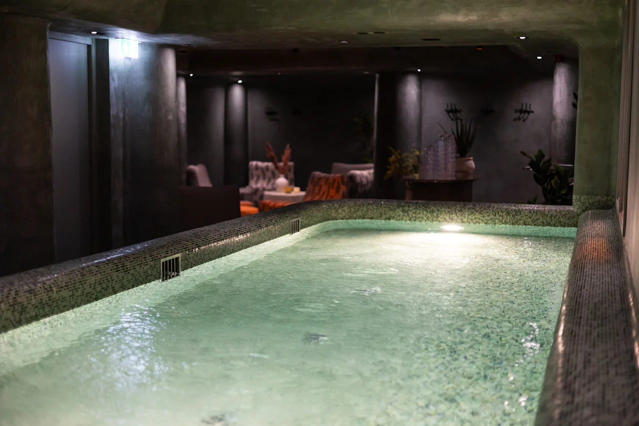 Rauhallinen sisäuima-allas tai poreallas vihreillä laatoilla, jota ympäröi hämärästi valaistu oleskelutila kasveineen, luoden rentouttavan kylpylätunnelman.