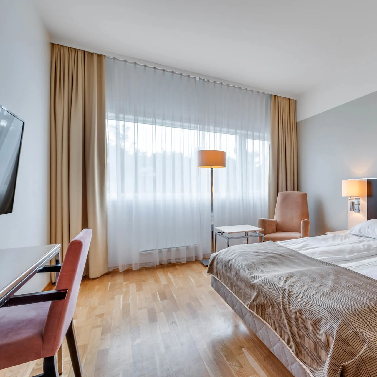 A tidy hotel room with a large bed, armchair, and a table under ambient lighting near a curtained window.