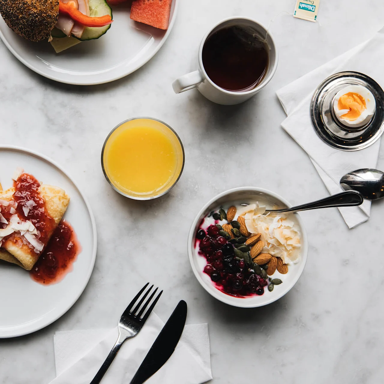 Pancakes topped with strawberry sauce and coconut flakes, served on a white plate next to yogurt with berries and nuts, orange juice, tea, and a boiled egg on a marble table.