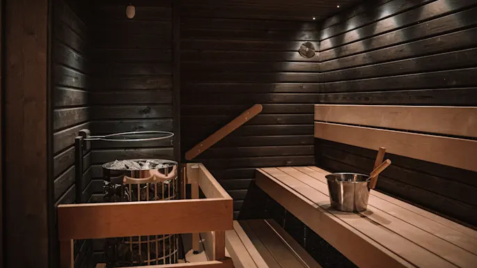 En trebadstue har benkesitteplasser og en metallovn med steiner. En metallbøtte og øse hviler på benken. Det svakt opplyste rommet har trevegger og tak.