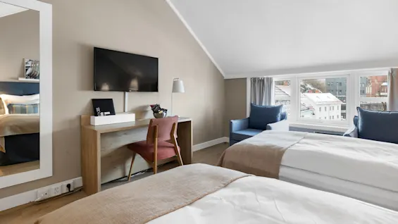 Et hotellrom med to senger, et skrivebord med stol, en flatskjerm-TV på veggen, og store vinduer med utsikt over et snødekket bylandskap.