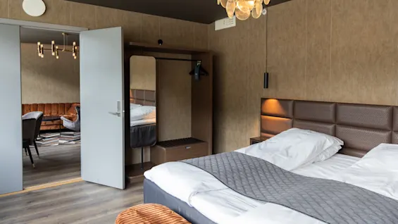 En prydligt bäddad säng med grått sänglinne står i ett modernt hotellrum, med bruna väggar och designerbelysning. En öppen dörr visar ett stilfullt sällskapsrum med grå stolar.