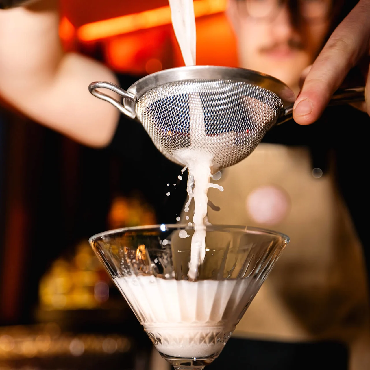 En cocktail silas genom en metallnät till ett glas, hålls av en bartender. Bakgrunden är en svagt upplyst bar med varm belysning.
