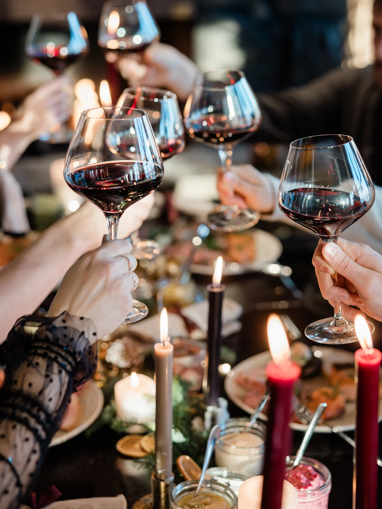 Glas rött vin skålas, hållna av flera händer runt ett bord dekorerat med ljus och mat, i en varmt upplyst, mysig miljö.