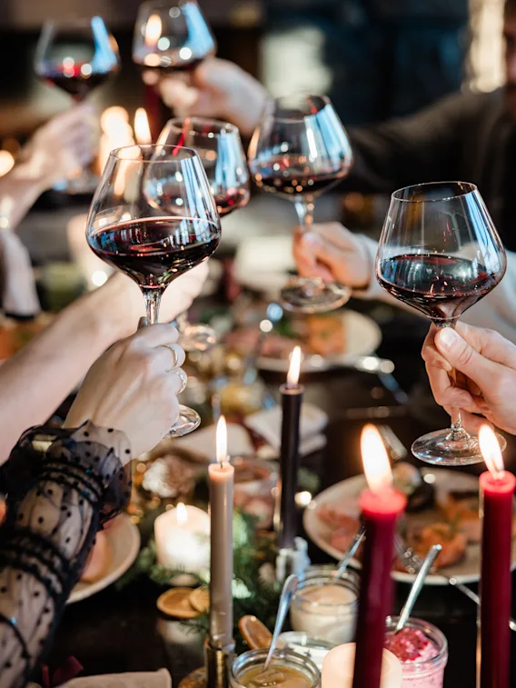 Glas rött vin skålas, hållna av flera händer runt ett bord dekorerat med ljus och mat, i en varmt upplyst, mysig miljö.