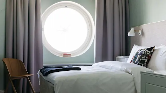 En seng med et mønstret pudebetræk står ved et rundt vindue med tykke grå gardiner. En træstol og puf fuldender det minimalistiske rum.