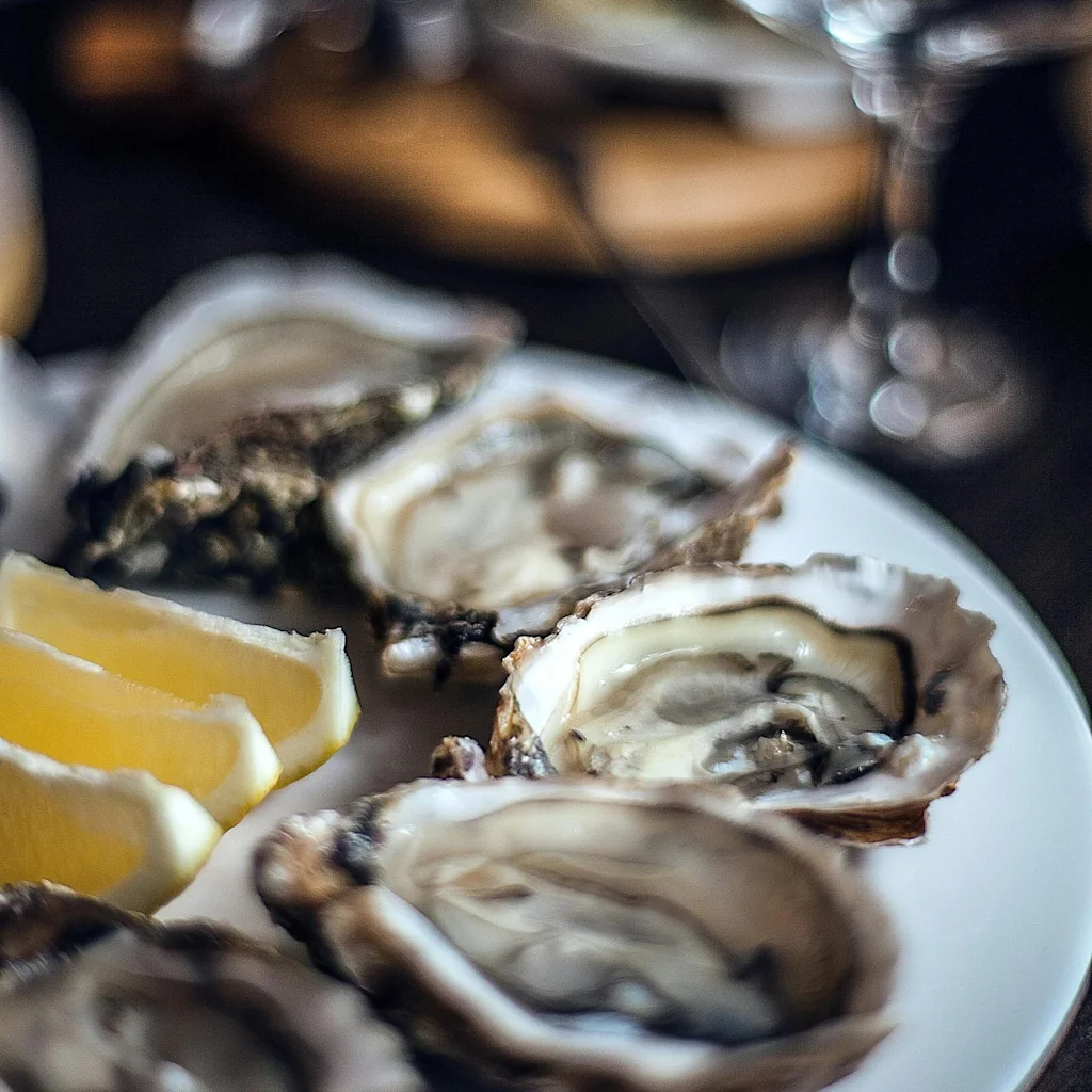 En tallerken med friske østers og citronbåde, klar til at blive nydt, med et sløret glas i baggrunden.