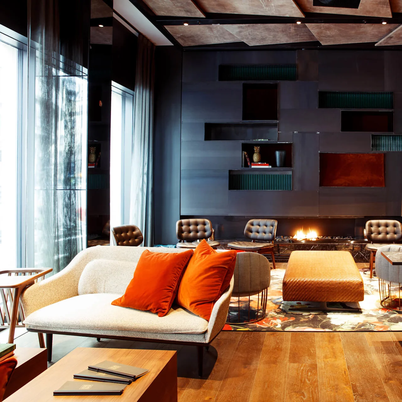 A contemporary lounge featuring a mix of chairs and sofas around a central fireplace. The space is decorated with modern art, lush plants, and ambient lighting.