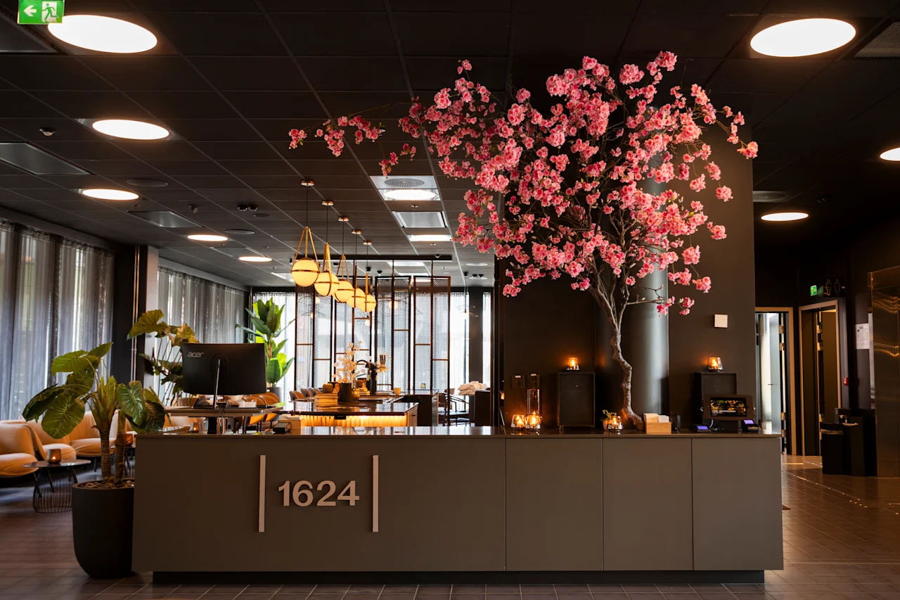 En moderne hotellobby med en resepsjon som viser '1624', et stort rosa kirsebærtre og stilig belysning.