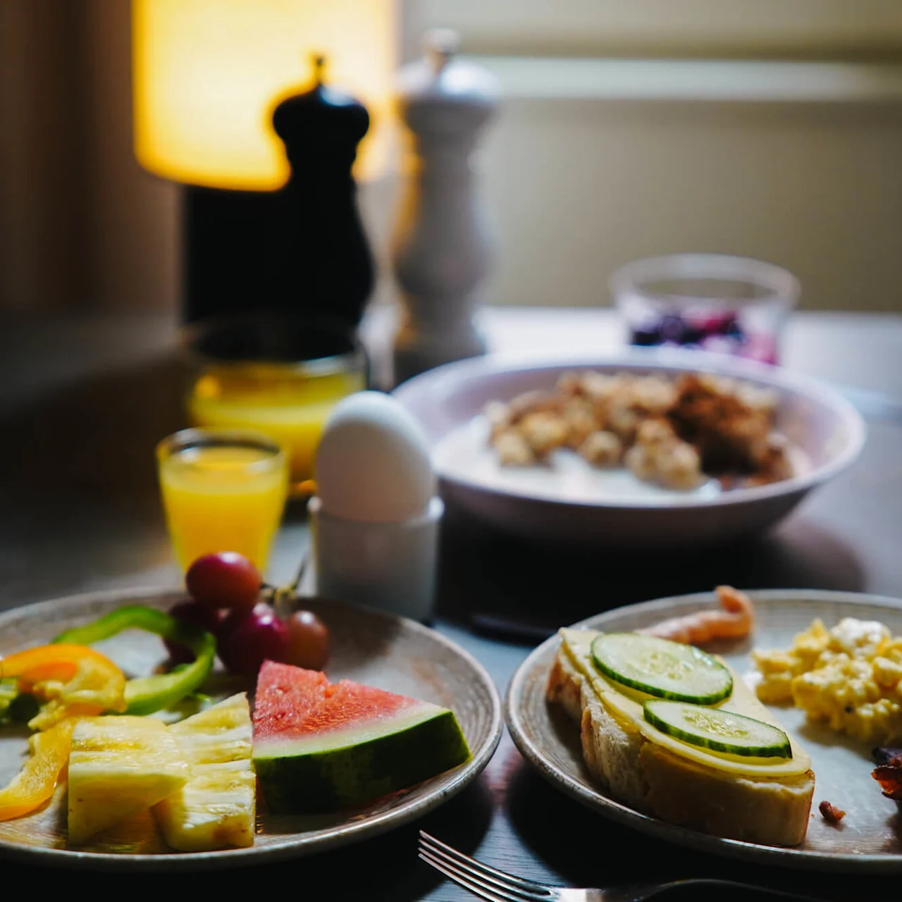 Tallrikar med skivad frukt, grönsaker, rostat bröd och äggröra på ett bord; ett kokt ägg i hållare och ett glas apelsinjuice bredvid. Mörkt belyst matsal.