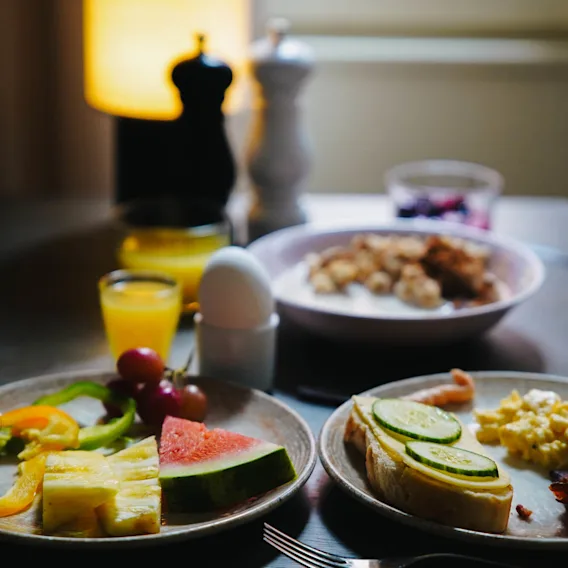 Tallrikar med skivad frukt, grönsaker, rostat bröd och äggröra på ett bord; ett kokt ägg i hållare och ett glas apelsinjuice bredvid. Mörkt belyst matsal.