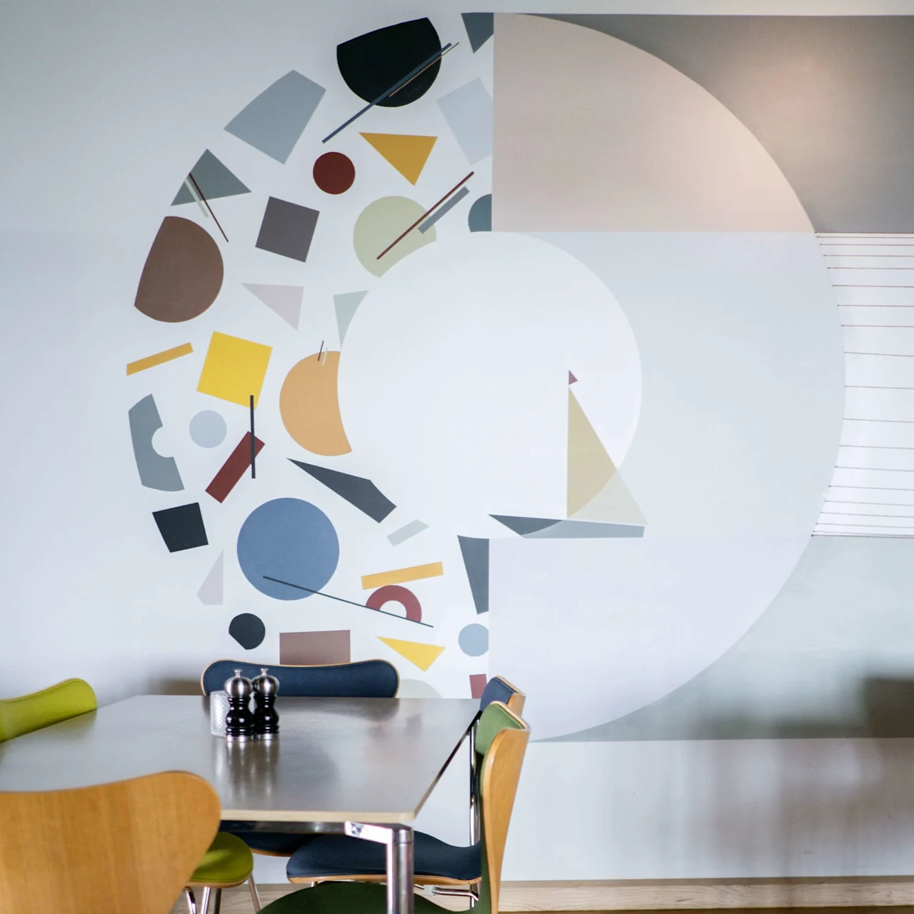 Matbord med färgglada stolar omkring, står på ett trägolv bredvid en abstrakt mural av geometriska former och en segelbåt på väggen. Matta med varma färger.