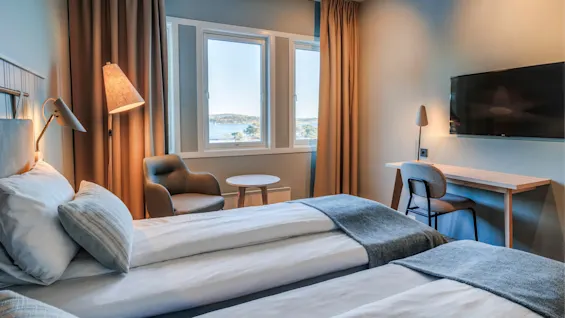 Two neatly made beds with gray throws rest in a bright hotel room featuring a cozy armchair, a small table, and a desk with a lamp. Large windows show a scenic outdoor view.