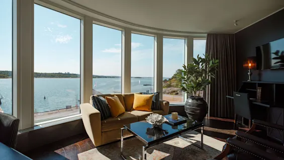 En moderne stue med en buet vinduesvæg, der tilbyder panoramaudsigt over havet og en klippefyldt kystlinje.