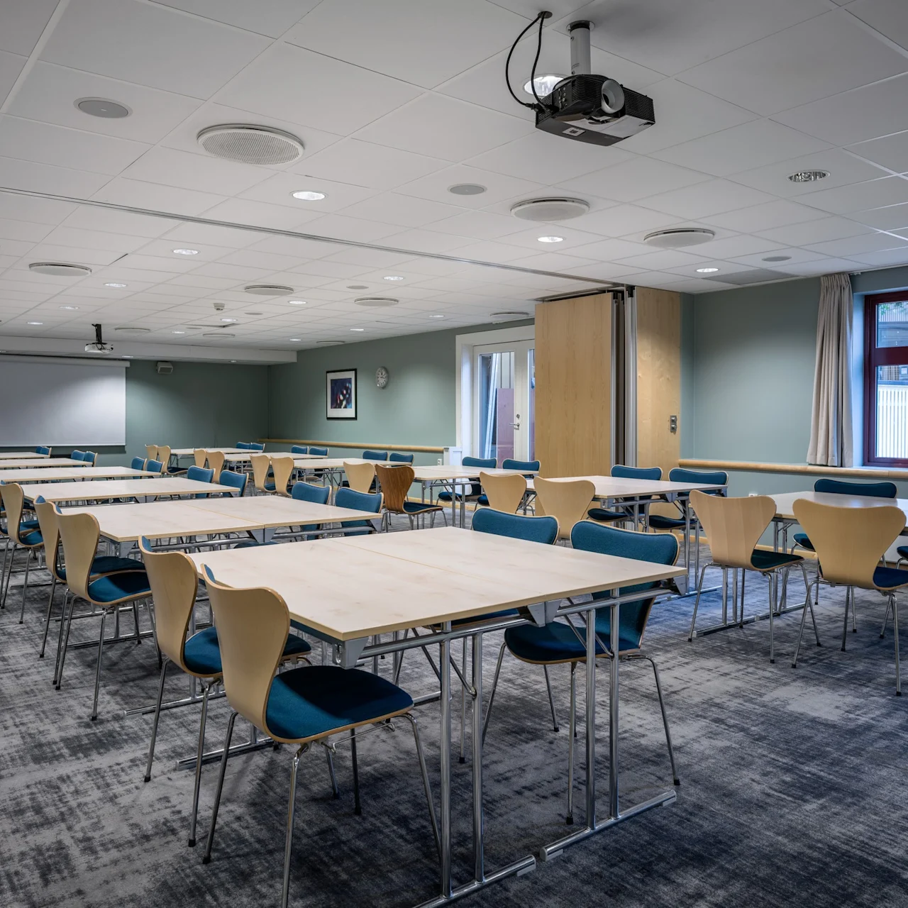 Bord och stolar är ordnade i rader i ett konferensrum med en projektorduk. Rummet har ljusblå väggar, en grå matta och stora fönster som släpper in naturligt ljus.