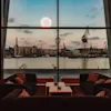 En moderne lounge med mørke sofaer og mønstrede puder vender mod et stort vindue, der viser en havneudsigt med bygninger, kraner og en solnedgangssky, mens fyrværkeri lyser op horisonten.