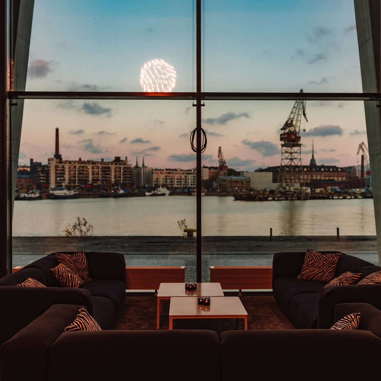En moderne lounge med mørke sofaer og mønstrede puder vender mod et stort vindue, der viser en havneudsigt med bygninger, kraner og en solnedgangssky, mens fyrværkeri lyser op horisonten.