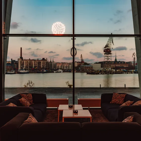 Moderni lounge tummilla sohvilla ja kuvioiduilla tyynyillä kohtaa suuren ikkunan, josta avautuu satamanäkymä rakennuksineen, nostureineen ja auringonlaskutaivainneen, samalla kun ilotulitteet valaisevat horisontin.