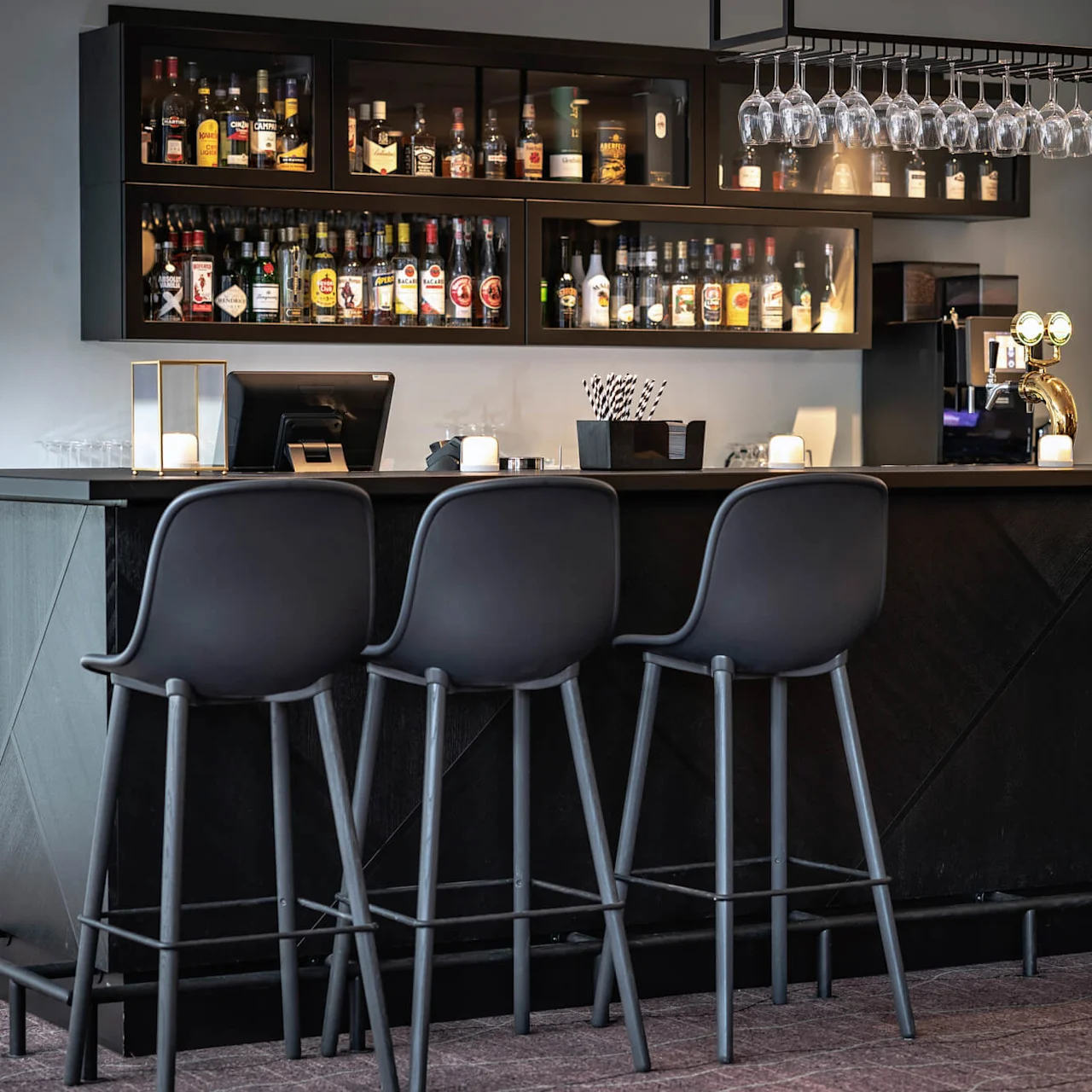 En elegant bar med høje stole vender mod hylder med pænt arrangerede spiritusflasker. Glas hænger ovenover, og blød belysning skaber en hyggelig atmosfære.