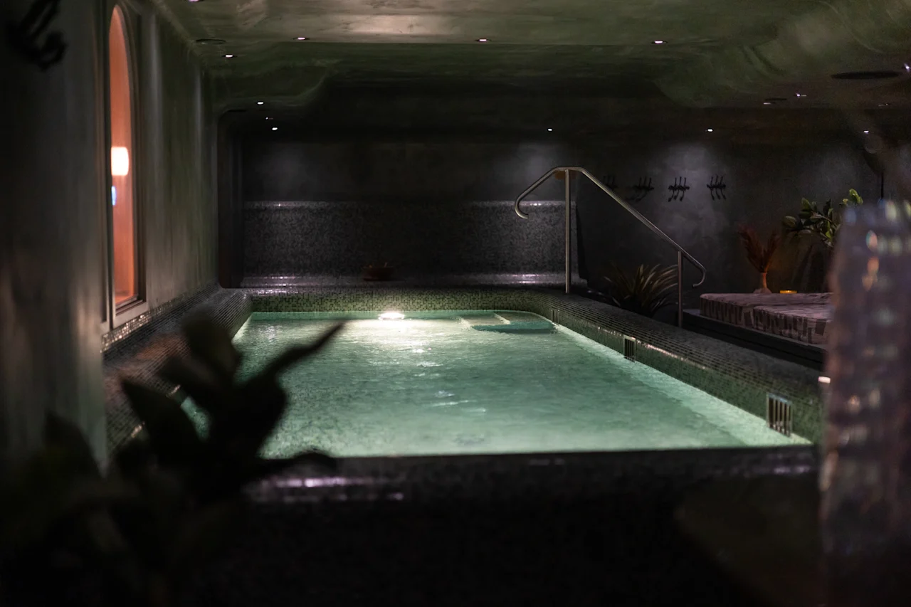 Hämärästi valaistu sisäuima-allasalue rauhallisella tunnelmalla, jossa on kimalteleva uima-allas, kaide ja hienovaraisia kasvikoristeita.
